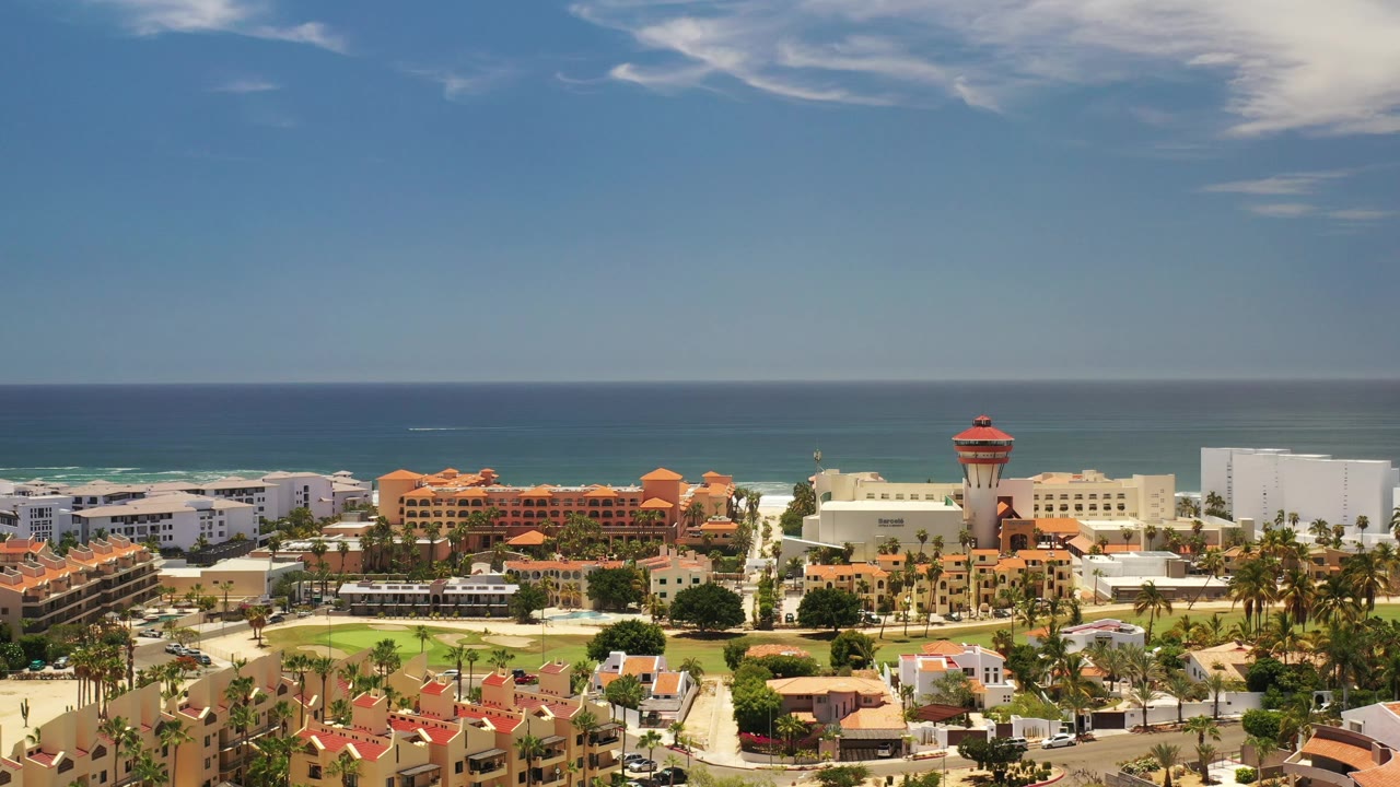 Aerial frame over Los Cabos showing the ocean horizon higher in the composition, resort-area buildings, a central green courtyard, palm trees, and no visible people.