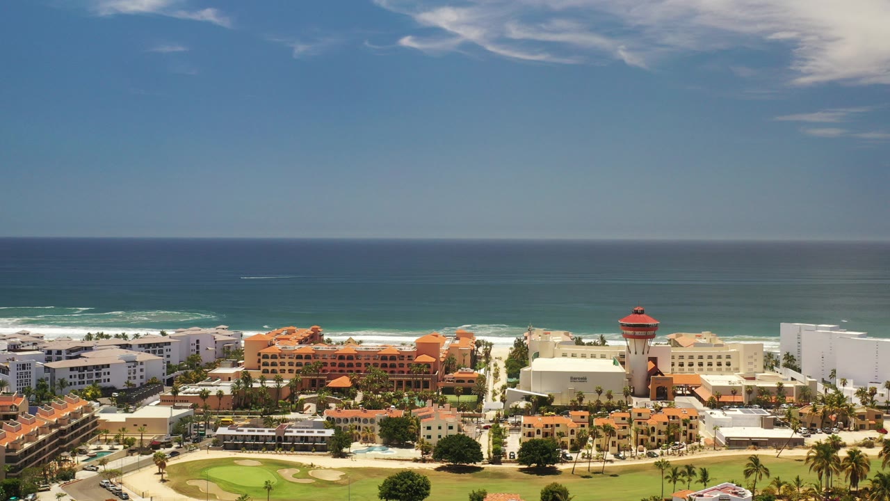 Wide coastal view with ocean, surf, resort buildings, palms, and blue sky.