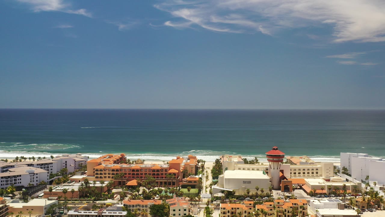 Opening aerial frame over Los Cabos, Mexico, with terracotta and white buildings below, surf beyond the beach, open ocean on the horizon, and no visible people.