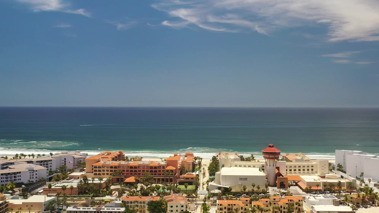 A wide Los Cabos coastal view shows beachfront hotels or apartment buildings, a red-roofed tower, white surf bands, blue ocean, and no visible people.