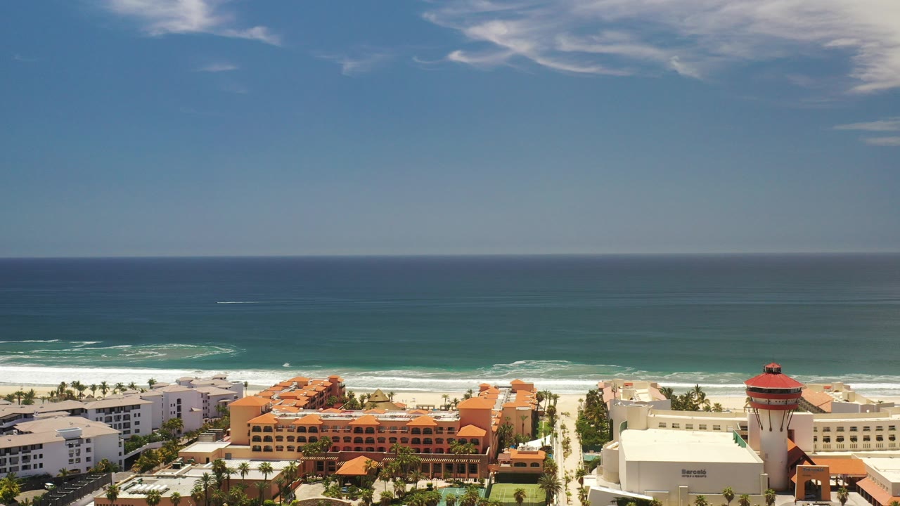 Opening aerial frame over Los Cabos, Mexico, with deep blue ocean beyond white surf, beachfront buildings along the lower edge, and no visible people.
