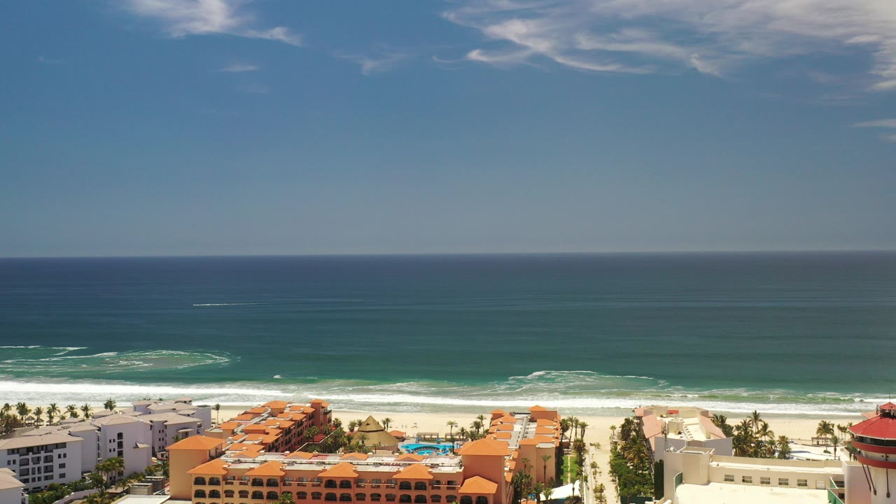 Opening frame of Los Cabos, Mexico from an elevated viewpoint, with orange-roofed beachfront buildings below, a pale beach, white surf, blue-green water, a straight horizon, and no visible people.