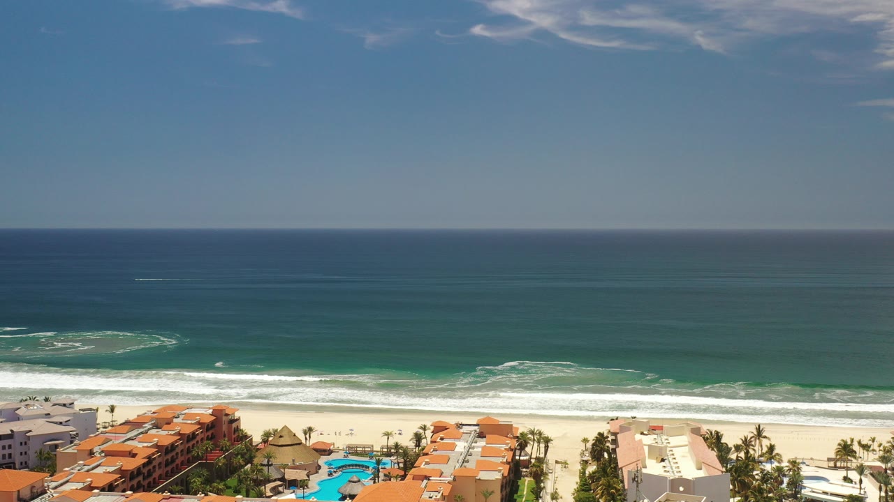 Aerial view of ocean waves, beach, resort roofs, palms, and pools.