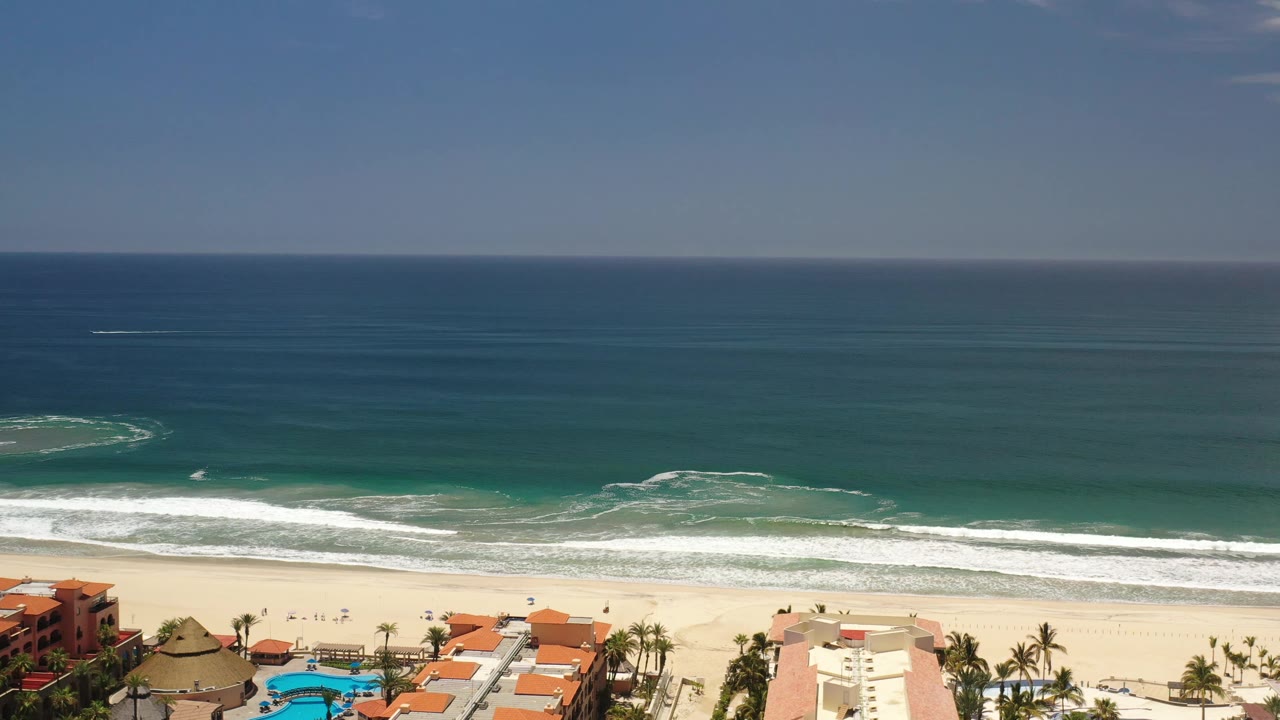 Aerial view in Los Cabos, Mexico, showing a long surf break across turquoise water with resort roofs, palms, and a pool at the bottom edge, no visible people.