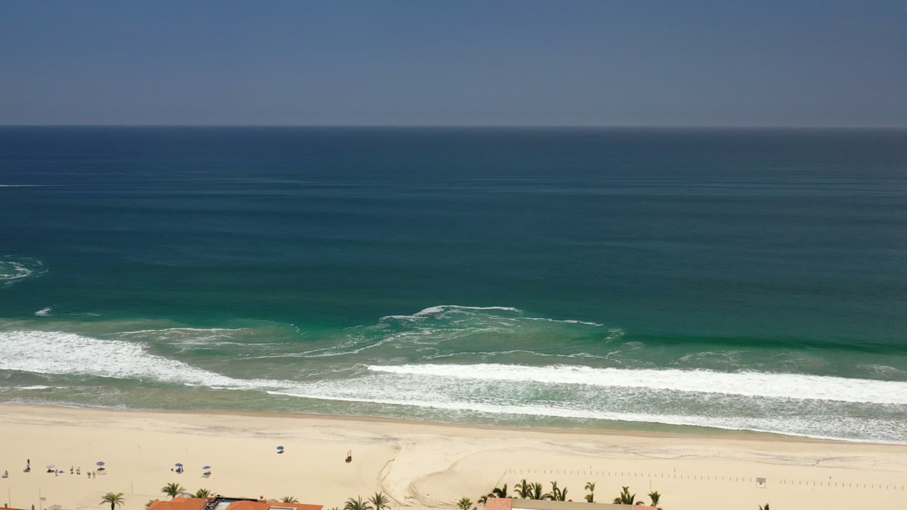 Los Cabos shoreline seen from a high angle, with a broad dark ocean horizon, green-blue nearshore water, curling white surf, and tiny distant figures on the sand.