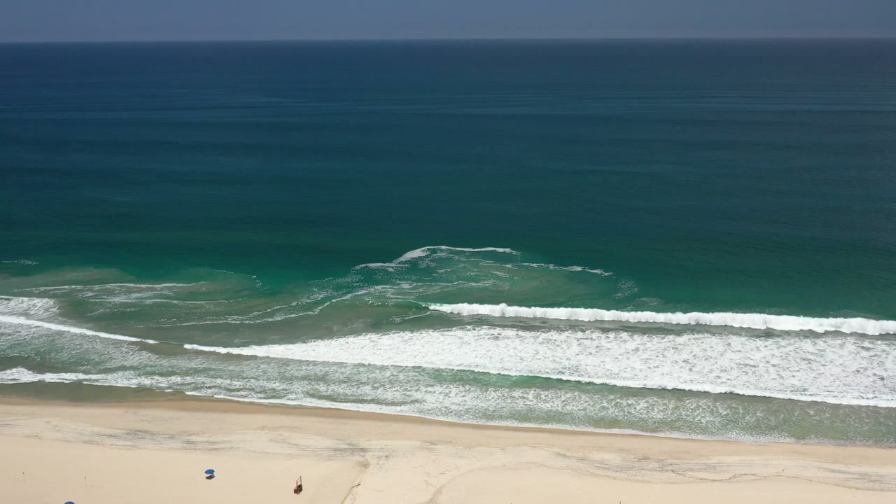 Aerial Los Cabos beach frame with a long white breaker crossing the middle, darker open ocean above it, pale sand below, and no clearly visible people in this moment.