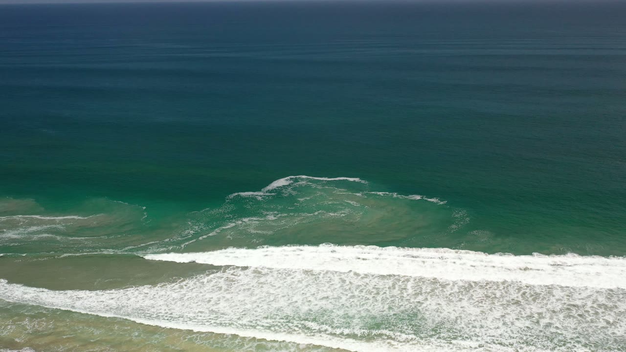Overhead view of green-blue surf breaking across sand in Los Cabos, Mexico, with open water above and no visible people.