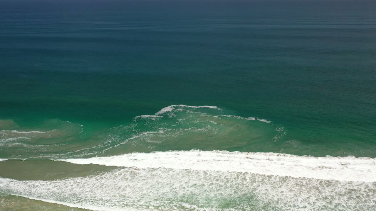 Aerial Los Cabos shoreline frame with a long white surf band over shallow sand and darker open water above, no visible people.