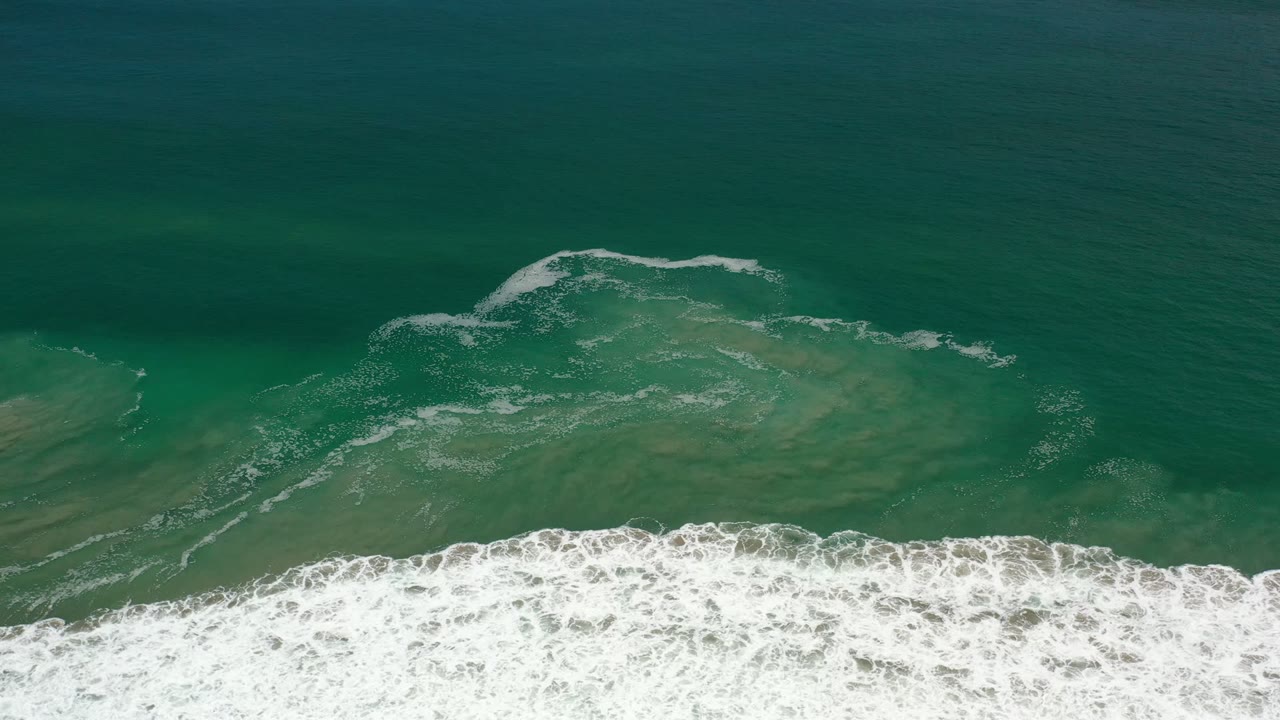 Overhead Los Cabos ocean frame with white foam curling across the lower third and darker green water filling the upper area, with no visible people.