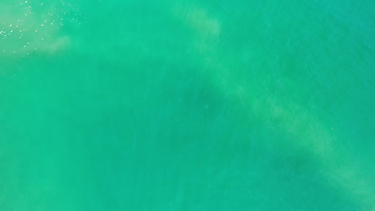Aerial frame of turquoise Los Cabos water with a soft cloudy foam patch left of center and scattered light specks, with no visible people.