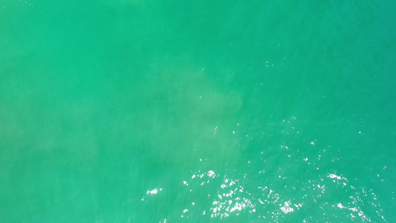 Overhead view of turquoise green ocean water in Los Cabos, Mexico, with scattered sun reflections near the lower left and no visible people.