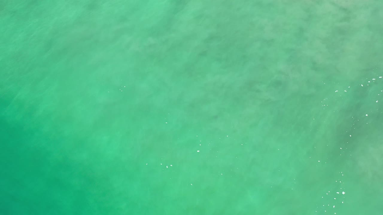 Overhead Los Cabos sea surface with green blue water, soft underwater patches, and a loose line of small sun glints toward the upper right, with no visible people.