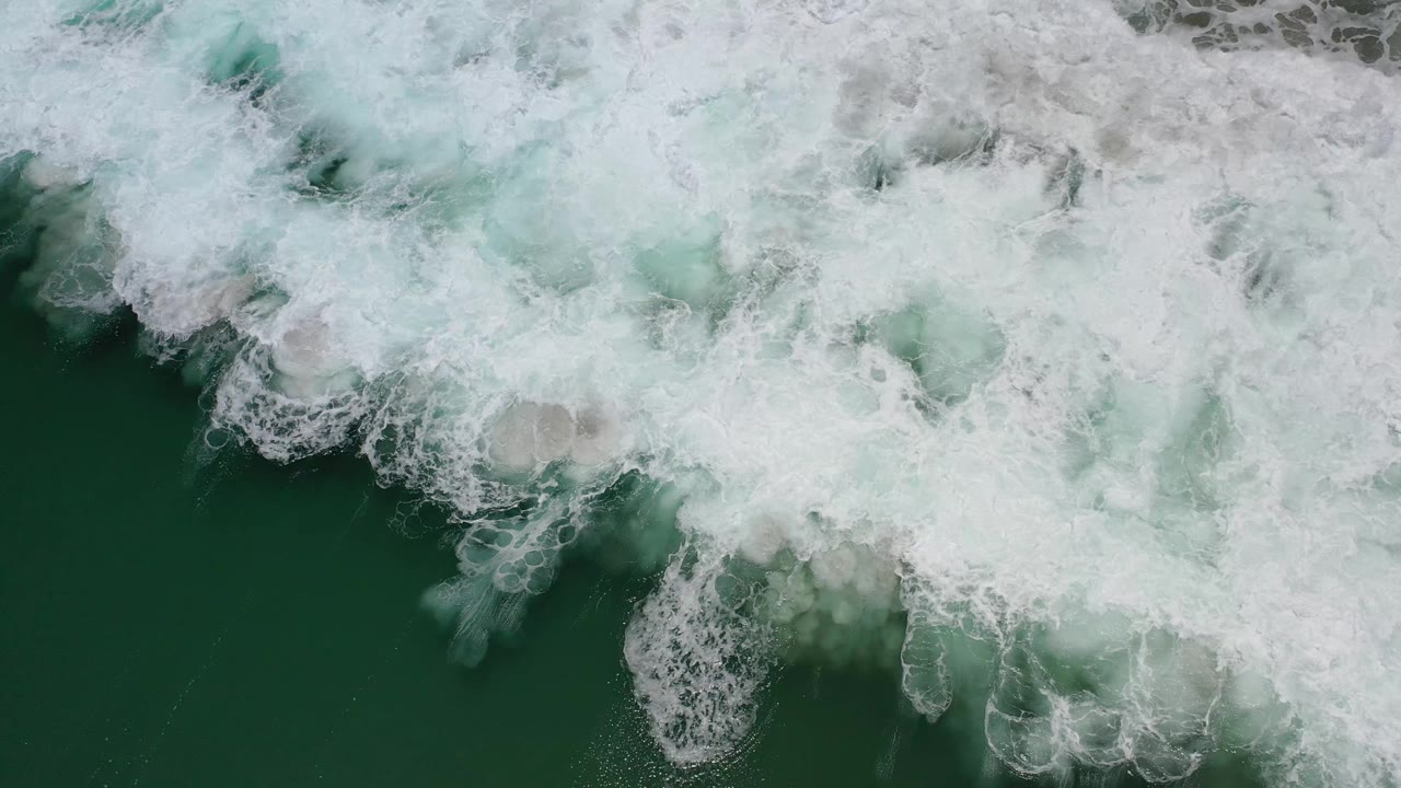 Aerial-style overhead frame of white ocean foam pushing farther across green water in Los Cabos, Mexico, with no visible people.