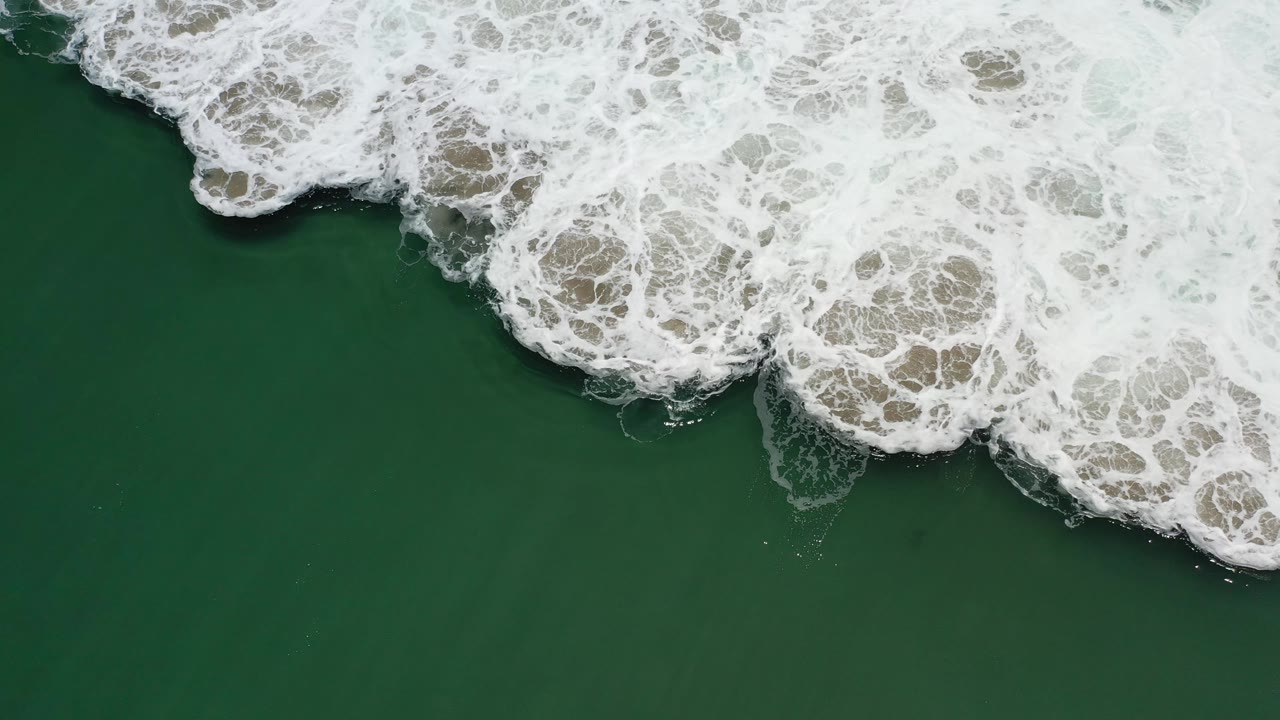 Aerial Los Cabos ocean frame with dark green water below and a broad band of white broken waves pushing across the upper right, no visible people.