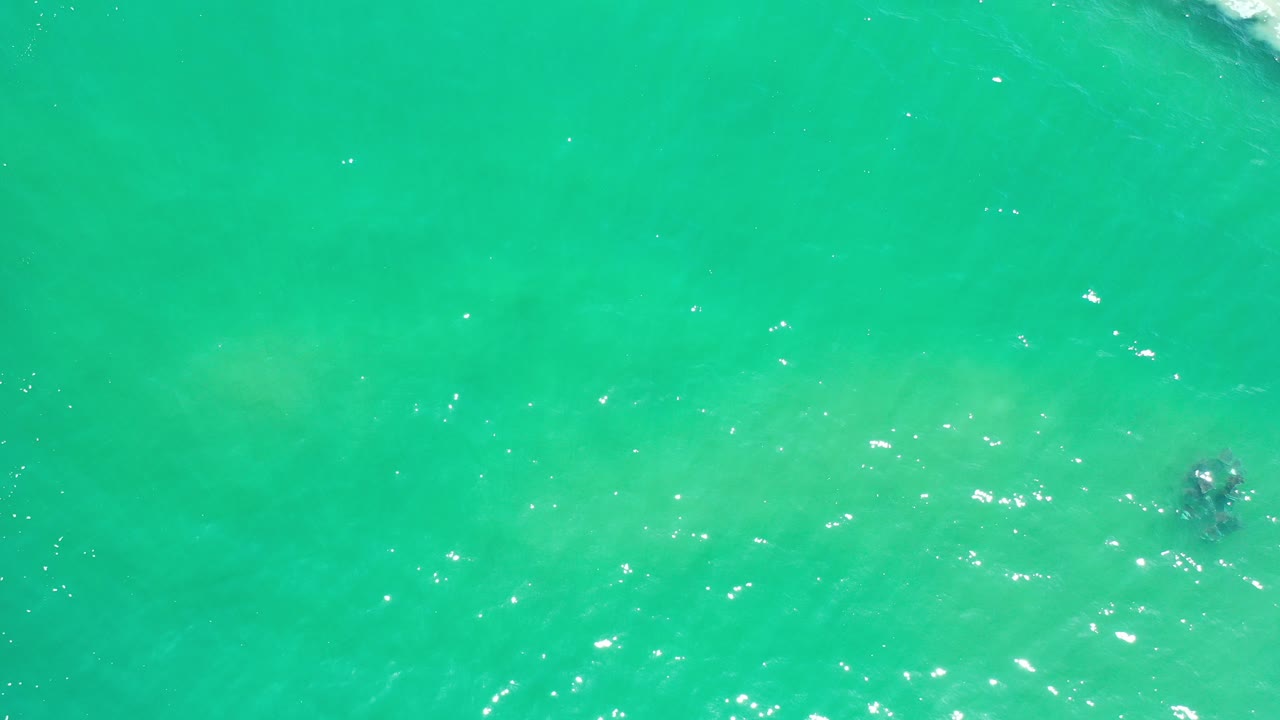Overhead frame of clear teal ocean water in Los Cabos, Mexico, with scattered sun reflections, faint darker areas below the surface, and no visible people.