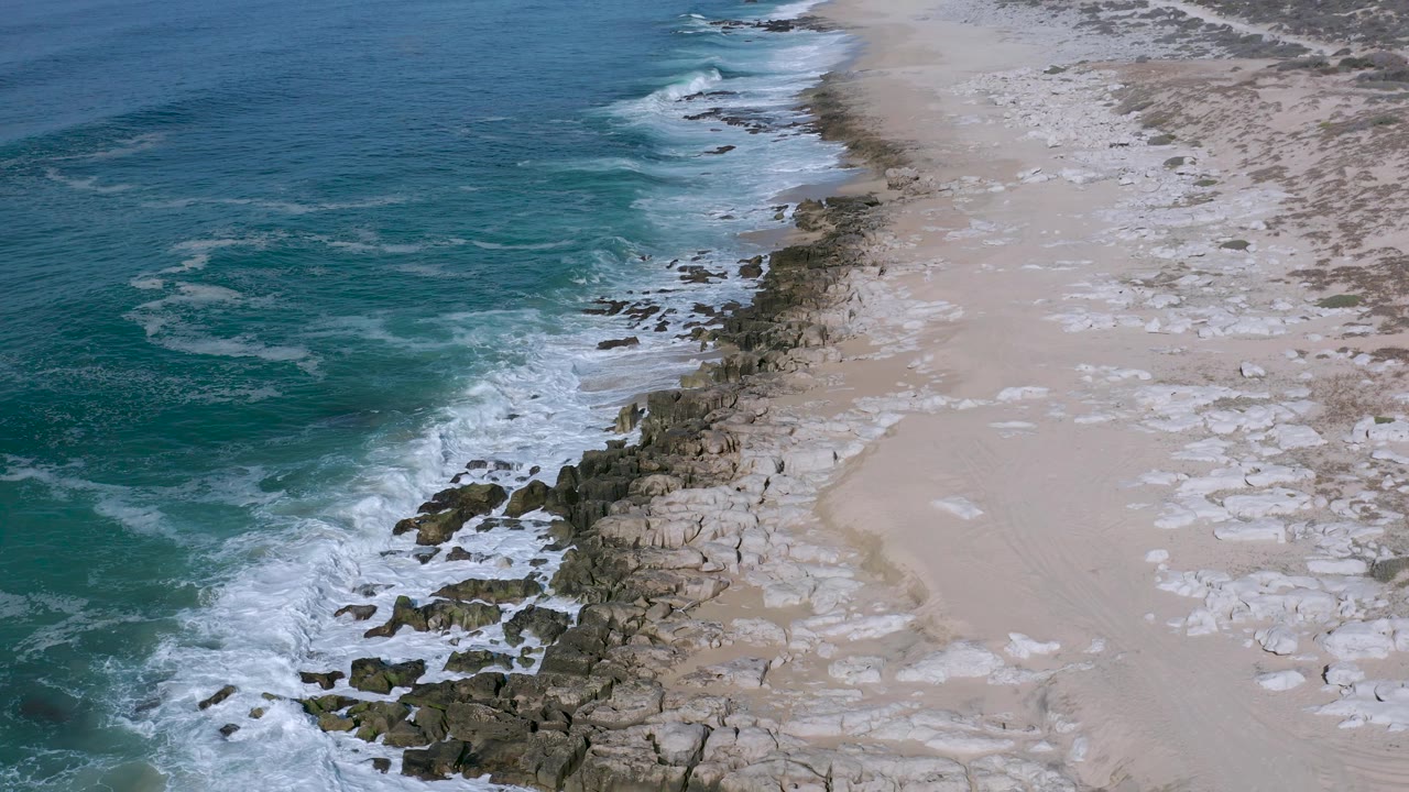 Aerial view of Los Cabos, Mexico shoreline with white water spreading across nearshore rocks and a broad pale beach area to the right, with no visible people.