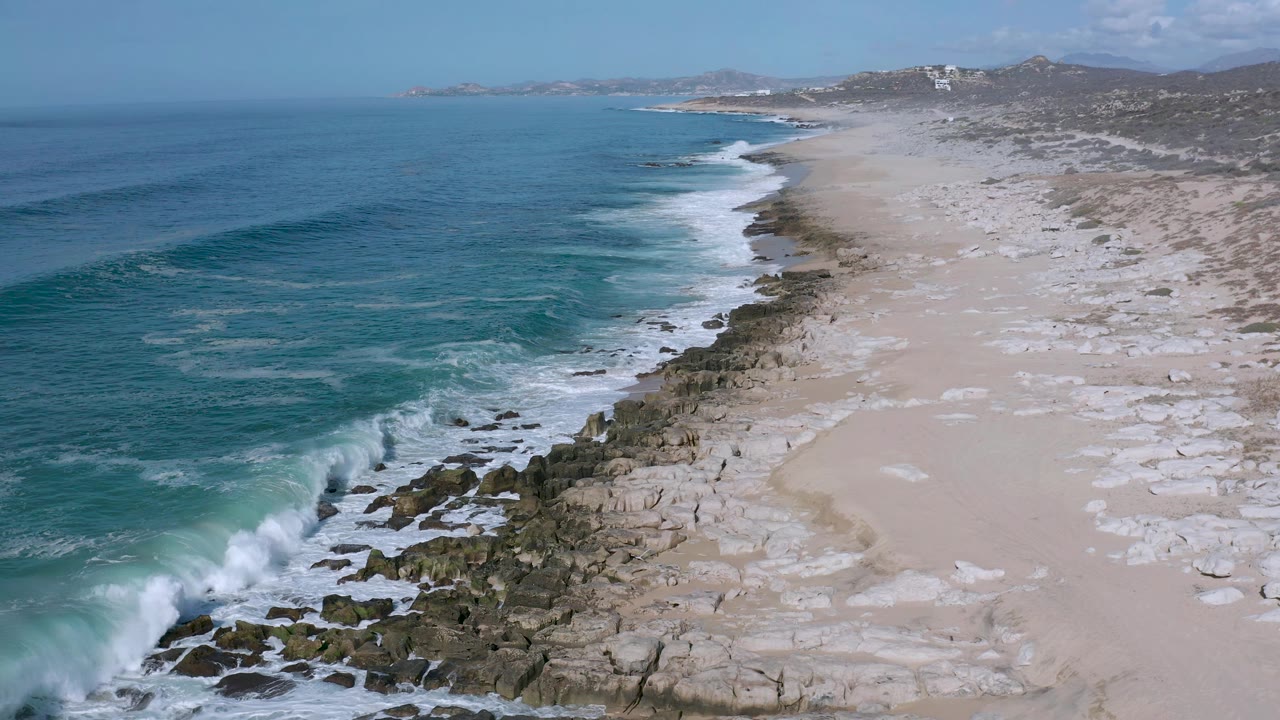Aerial Los Cabos shoreline view with a darker blue swell nearing the rocks, pale sand stretching inland, low hills beyond the beach, and no visible people.
