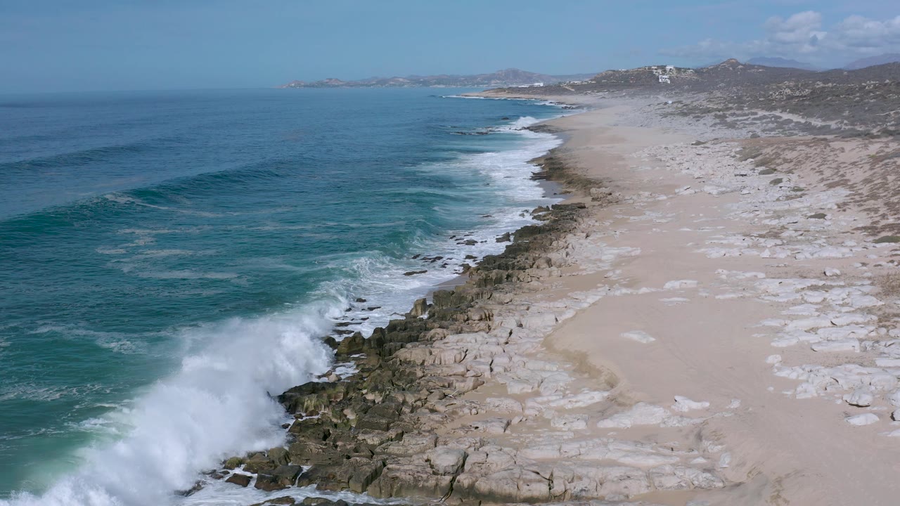 Aerial view of a wave breaking harder against the rocky edge of a Los Cabos beach, with white foam spreading along the shore and no visible people.
