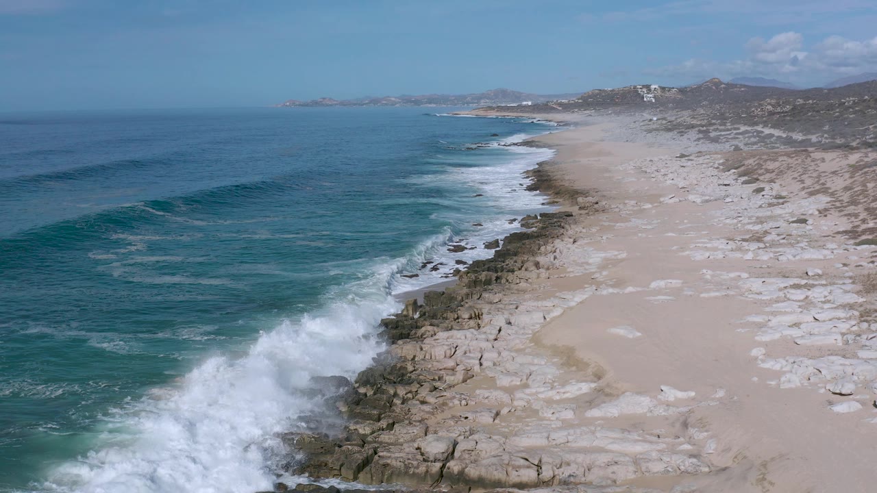 Aerial Los Cabos beach frame with white surf expanding over the rocks in the lower left, blue water offshore, pale sand to the right, and no visible people.