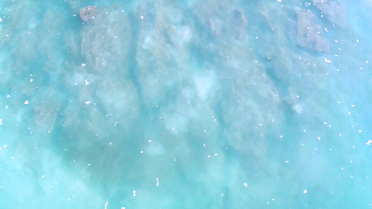 Top-down view of turquoise shallow ocean water over pale reef in Los Cabos, Mexico, with scattered white surface flecks and no visible people.