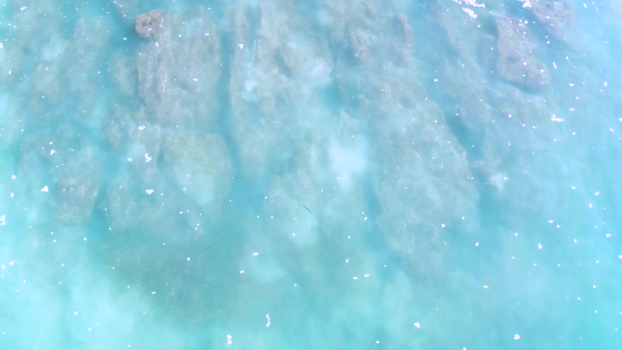 Clear turquoise ocean water reveals broad reef shapes below the surface in Los Cabos, Mexico, with bright specks of reflected light and no visible people.