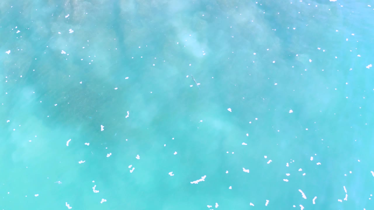 Overhead teal ocean view in Los Cabos with soft cloudy underwater patterns, scattered foam flecks, and no visible people.