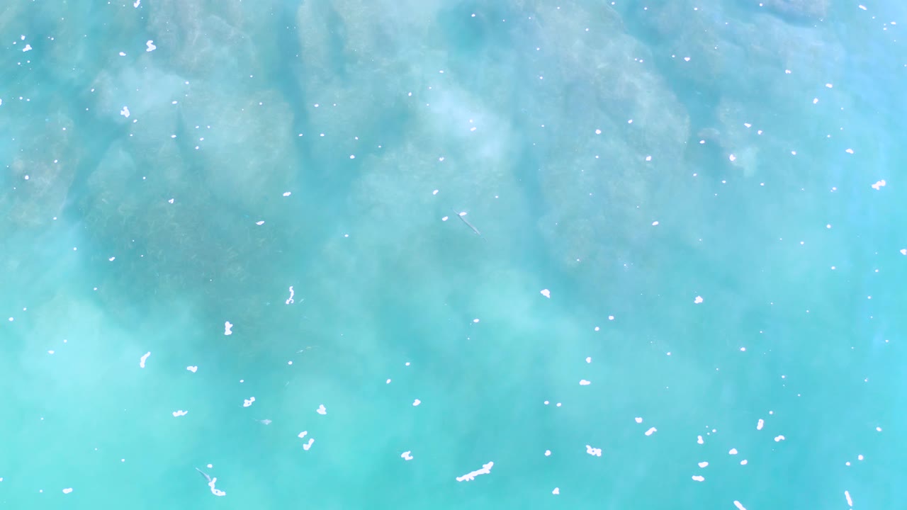 Top-down view of Los Cabos ocean water with pale white foam specks and soft teal patches under the surface, with no visible people.