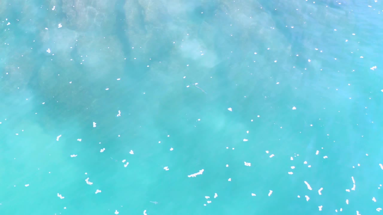 Mid-clip overhead frame of turquoise Los Cabos water with scattered white foam marks and darker blue patches under the surface, no visible people.