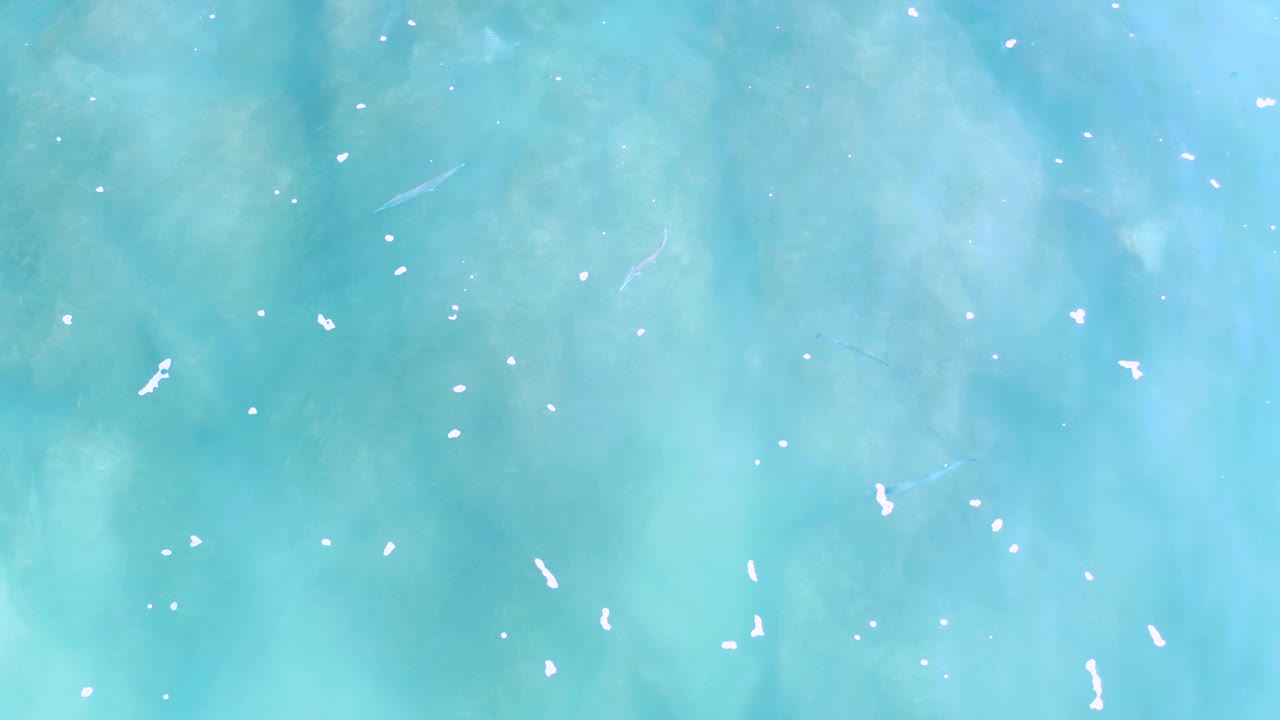 Overhead Los Cabos water view with denser foam flecks near the middle and muted underwater shadows beneath the turquoise surface, no visible people.