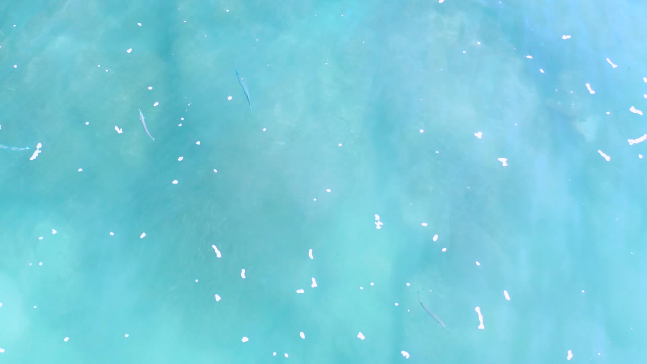 Overhead view of turquoise shallow water in Los Cabos, Mexico, with the bottom faintly visible, small white foam flecks across the surface, and no visible people.