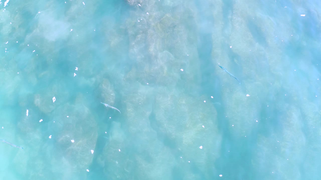Overhead aqua ocean surface with cloudy streaks, small white flecks, slightly darker blue patches, and no visible people.