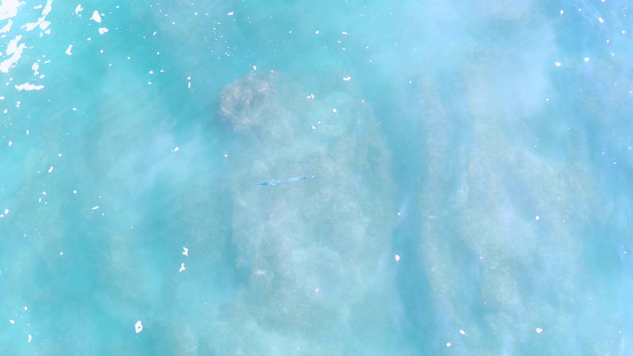 Overhead view of turquoise water in Los Cabos, Mexico, with pale submerged rock or sand forms, bright surface glints near the left edge, and no visible people.