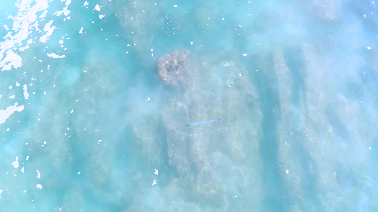 Overhead Los Cabos water view with a darker submerged patch near the upper left, clustered white glare along the left side, pale underwater texture, and no visible people.