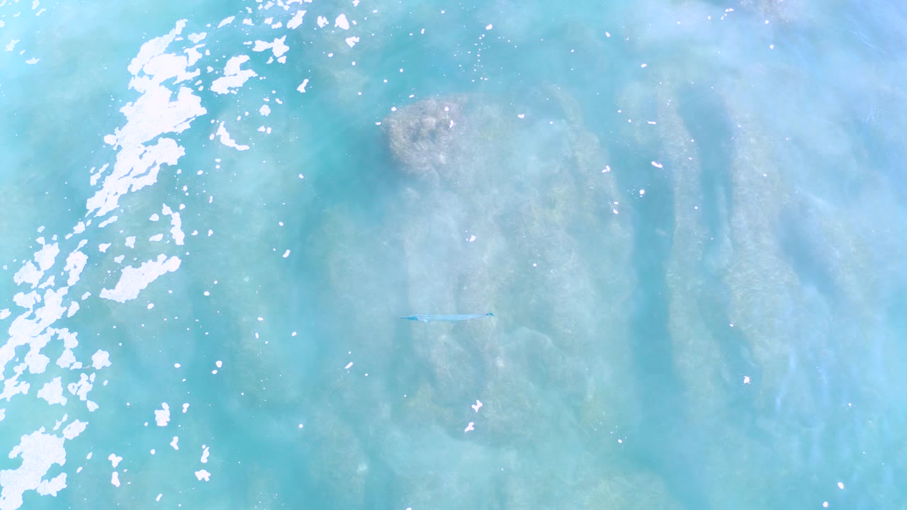 Overhead view in Los Cabos, Mexico, showing turquoise ocean water, white foam along the left side, submerged brown coral patches, and no visible people.