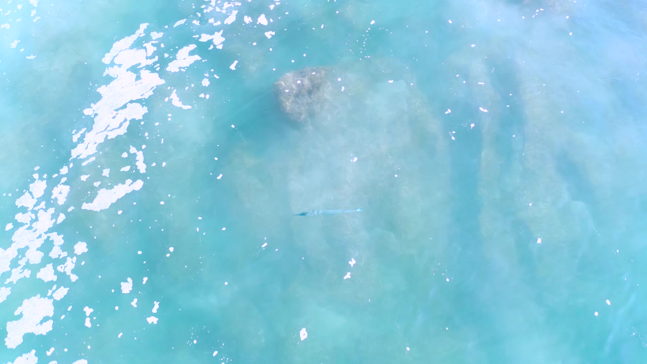 Overhead Los Cabos ocean view with white foam clustered near the left edge, scattered sun glints, a brown coral patch near the upper frame, and no visible people.