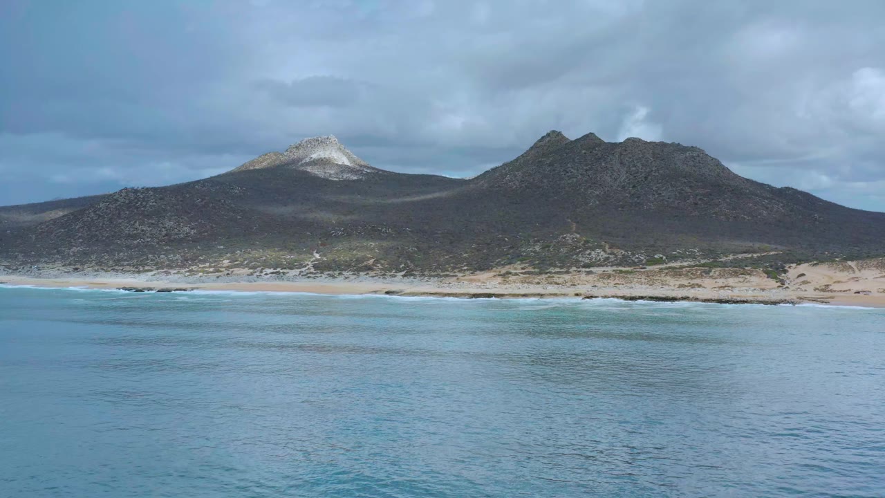 Los Cabos, Mexico ocean view with blue water in the foreground, a thin surf line, dark mountains, a white-topped peak at left, and no visible people.
