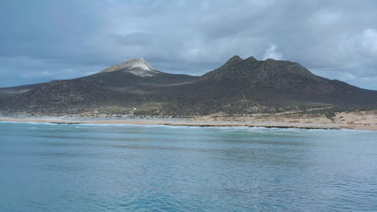 Los Cabos, Mexico ocean foreground with faint surface texture, a narrow beach band, dark rocky mountains, a bright white peak on the left side, and no visible people.