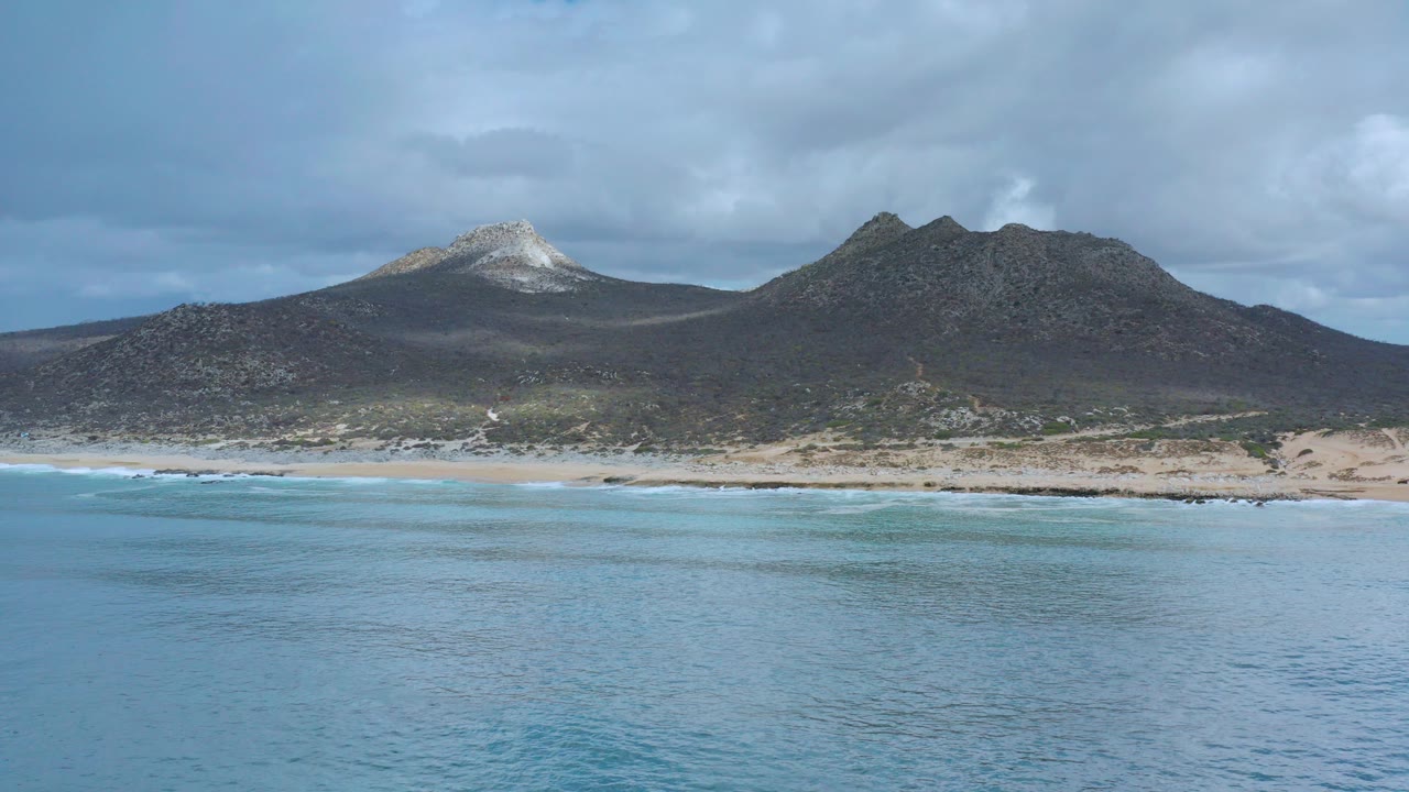 Wide coastal view in Los Cabos, Mexico, with turquoise water in front, a pale sandy beach, two mountain forms, heavy clouds, and no visible people.