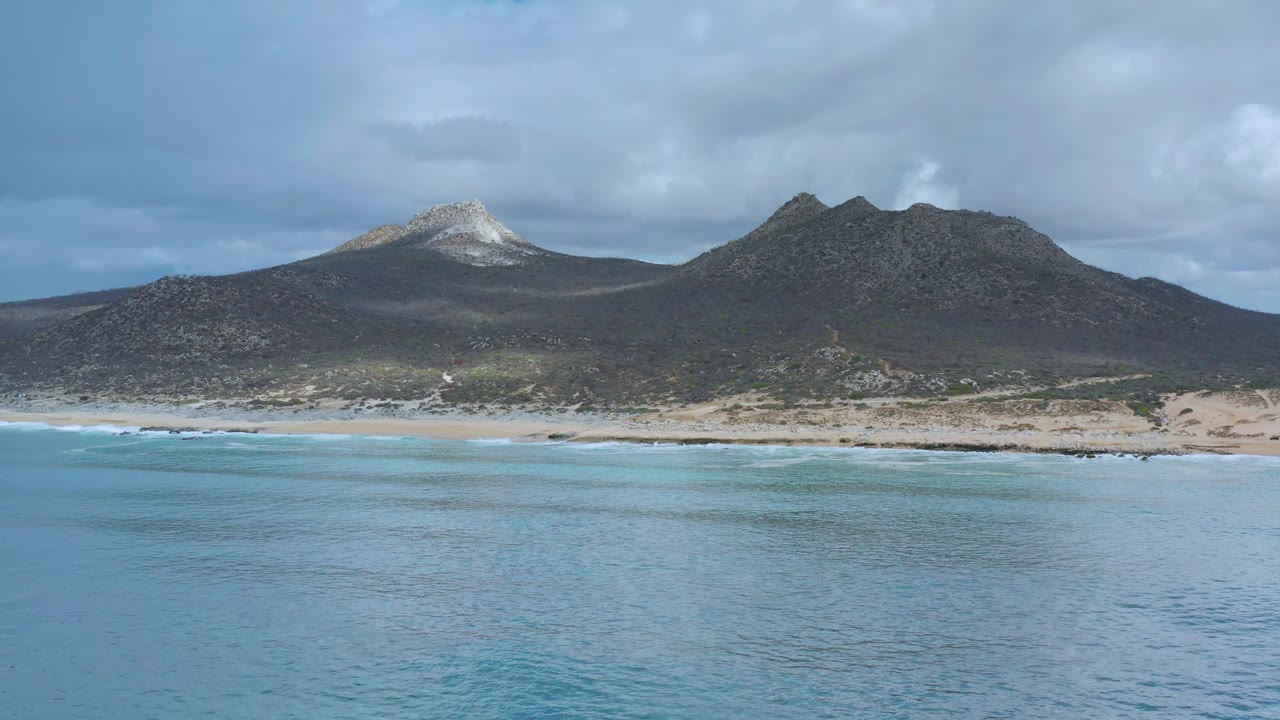 Los Cabos shoreline with rippling blue water in the foreground, a narrow sand strip, dark scrub at the mountain base, and no visible people.
