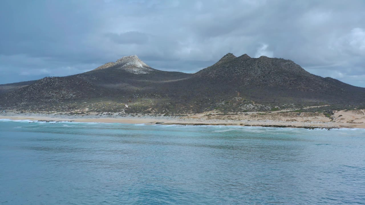 Wide Los Cabos beach frame with pale surf lines at the shore, turquoise water below, two dark mountain masses behind, and no visible people.