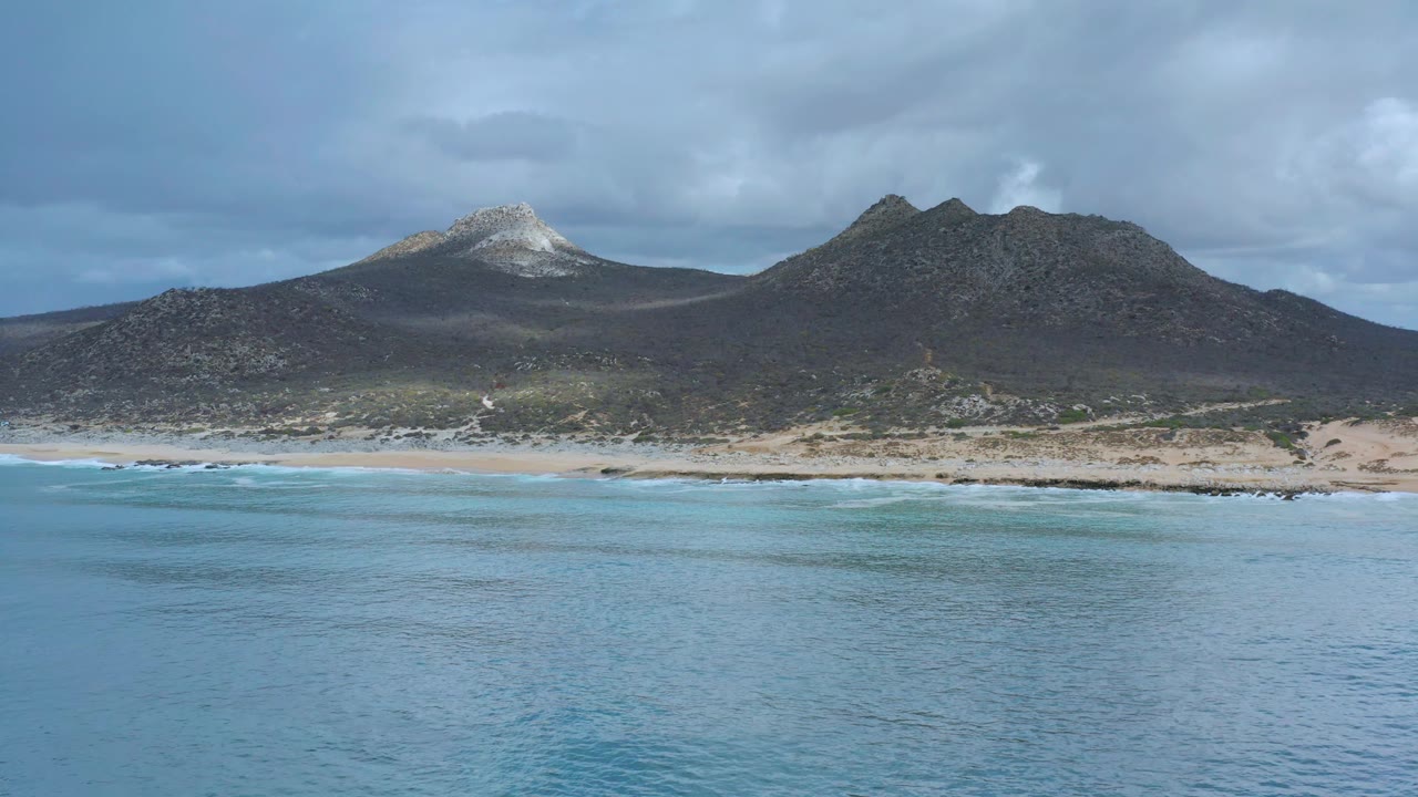 Los Cabos coastal landscape with an empty sandy beach across the center, dark mountains in the background, blue-gray clouds overhead, and no visible people.