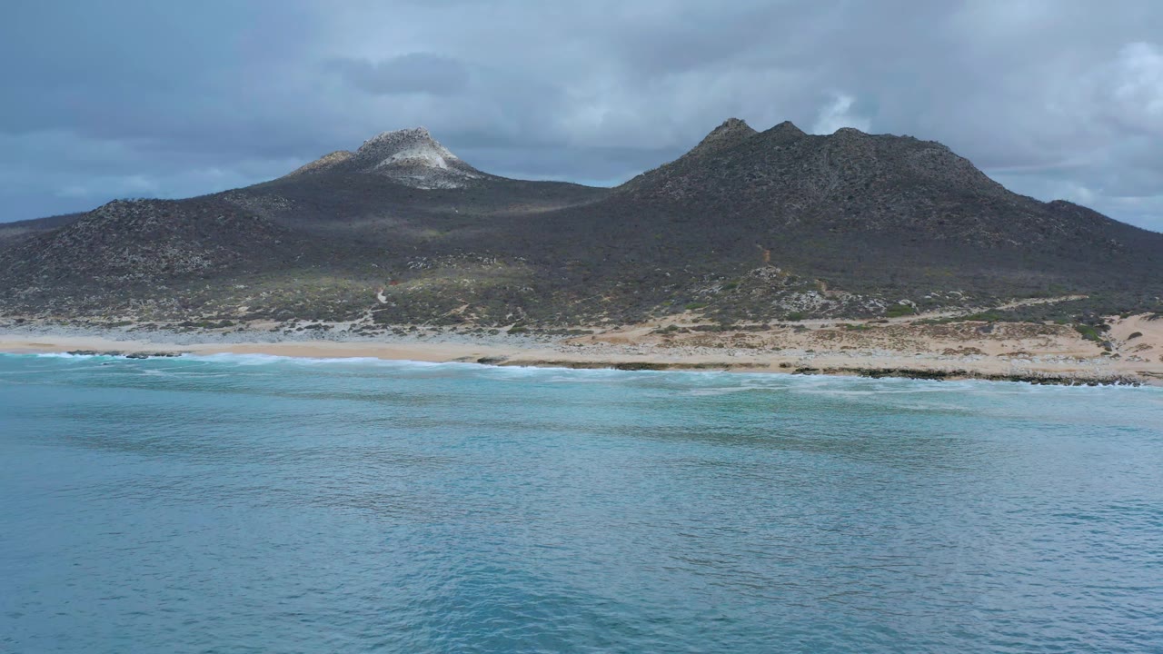 Aerial coastal view with turquoise water and dark hills under clouds.