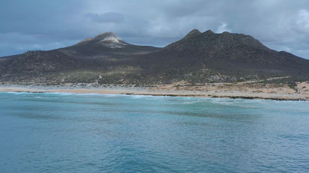 Los Cabos, Mexico wide coastline frame with rippled blue water, a narrow white surf edge, sandy beach, rugged dark ridge, cloudy sky, and no visible people.
