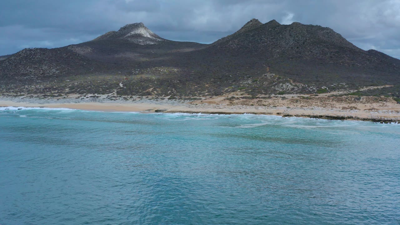 Wide aerial view of blue water, a sandy and rocky Los Cabos shoreline, dark mountains, cloud cover, and no visible people at the start of the clip.