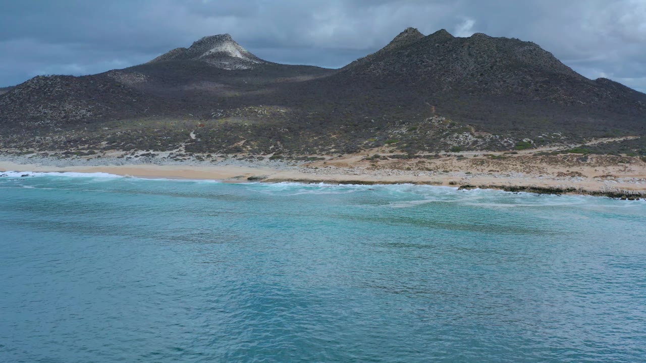 Wide Los Cabos coastal view with textured blue water in the foreground, a thin surf break on the beach, dark hills and mountain peaks beyond, and no visible people.