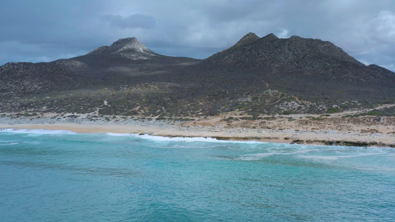 Opening aerial frame over the Los Cabos coast, with turquoise water in front, white surf on the beach, dark rocky hills behind, and no visible people.