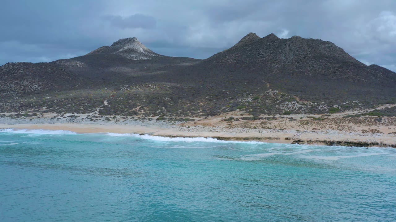Aerial view of the Los Cabos shoreline one second in, showing a broad band of turquoise sea, foamy waves, pale sand, scrubland, rocky mountains, and no visible people.