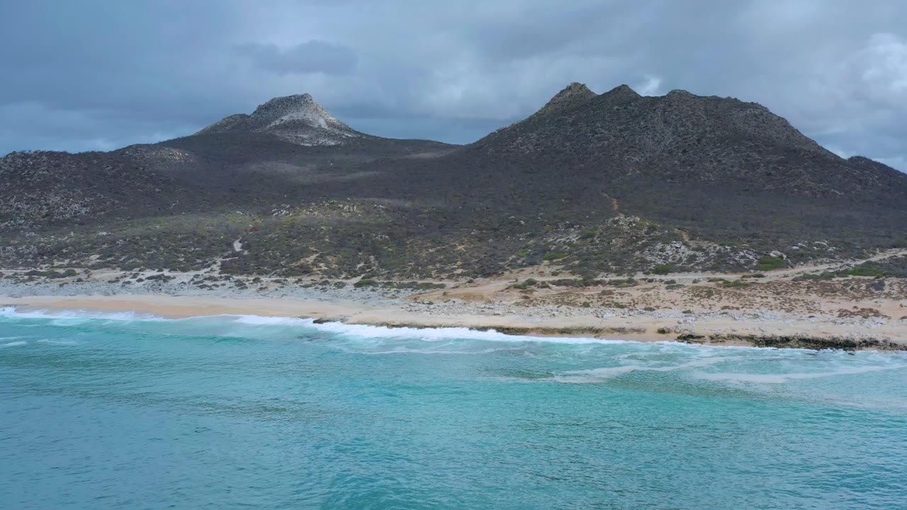 Aerial frame over Los Cabos with the waterline slightly lower in view, white wave bands crossing the shore, dark hills in the background, and no visible people.