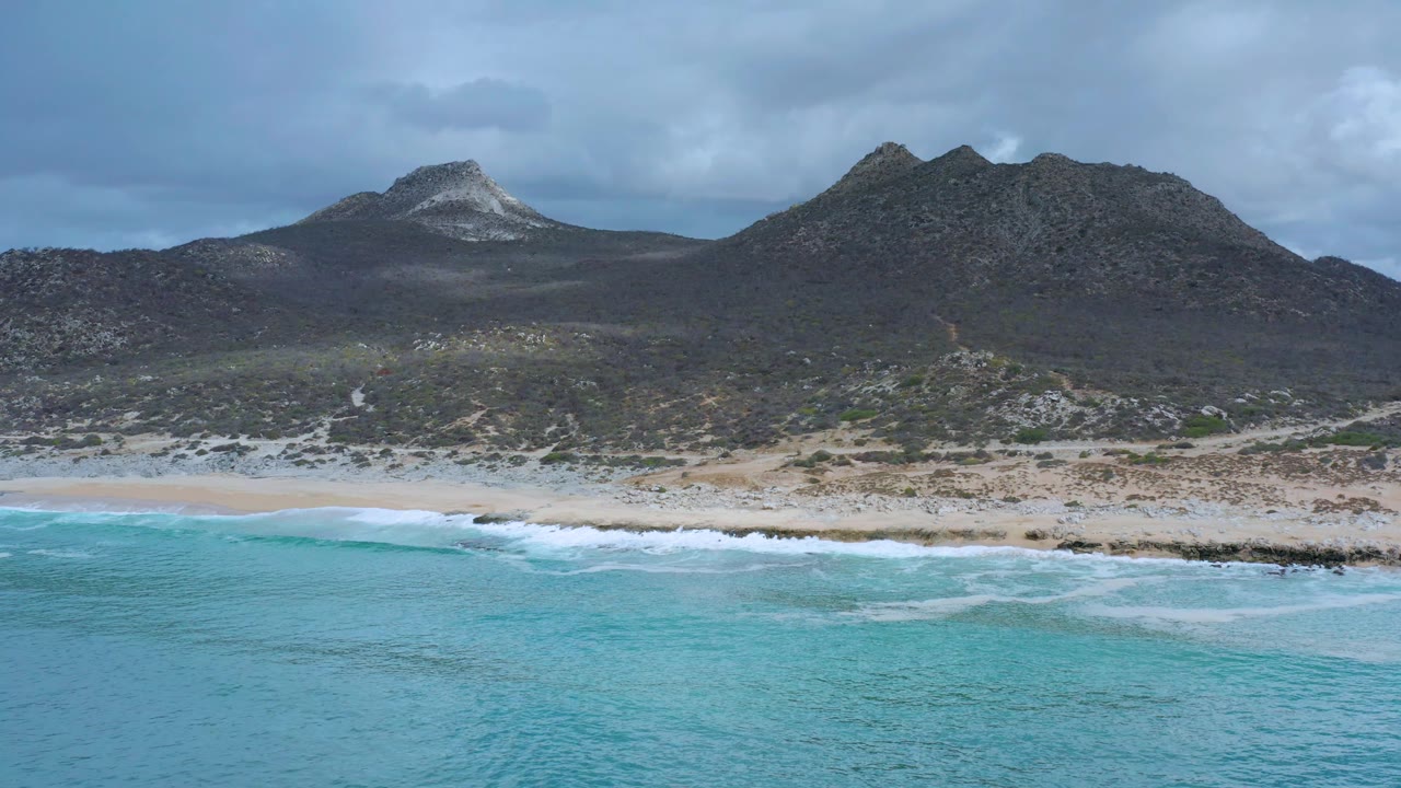 Mid-clip aerial view in Los Cabos, with more turquoise foreground water, a thin strip of breaking surf, pale beach, rocky scrub terrain, mountain slopes, and no visible people.