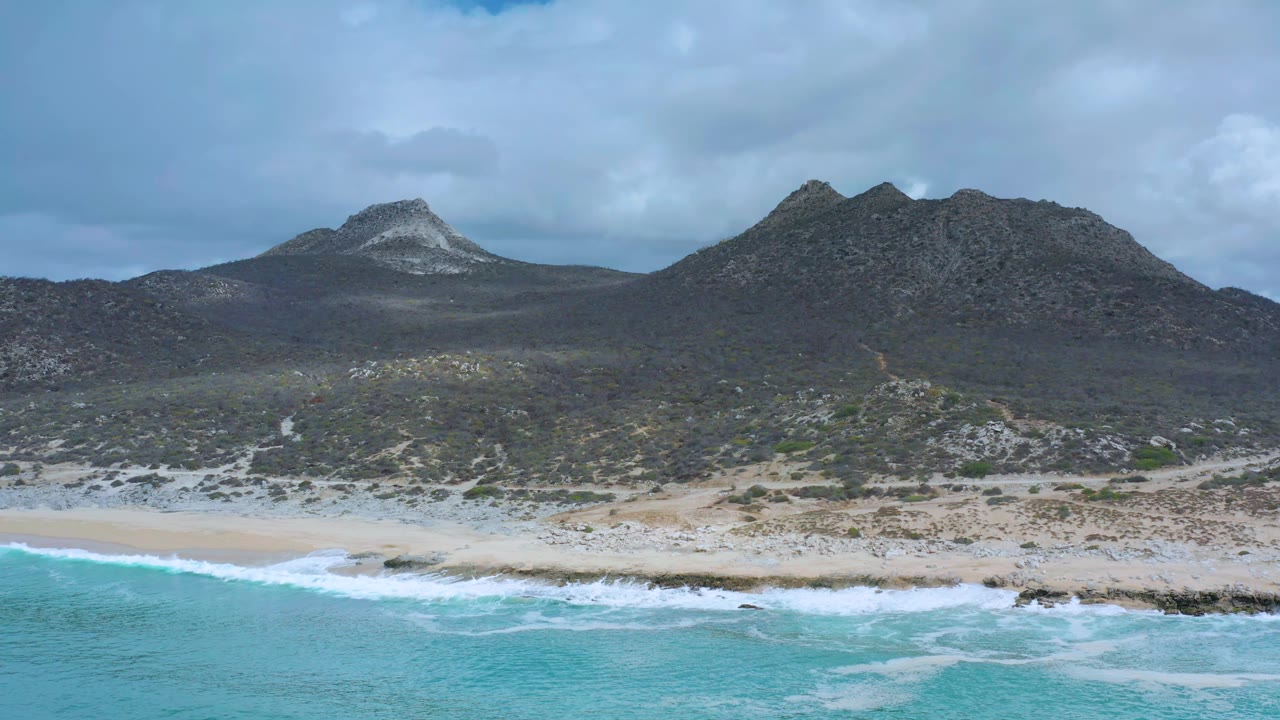 Los Cabos aerial frame with white waves crossing the lower edge, pale sand behind the surf, dark green scrubland, two rocky mountain forms, clouds overhead, and no visible people.